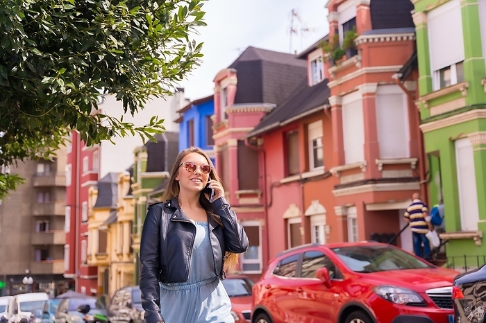 Young blonde woman in leather jacket walking talking on the phone, behind colorful facade