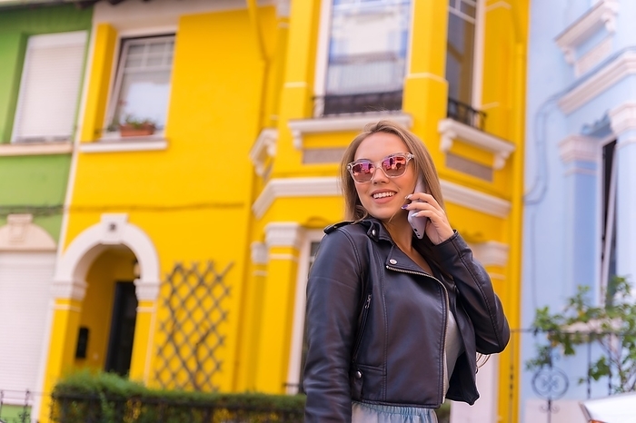Smiling young blonde woman in leather jacket talking on the phone, behind colorful facade