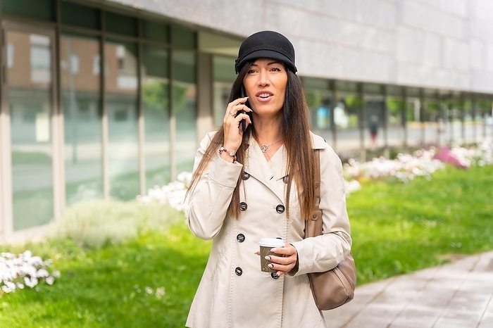 Businesswoman with a coffee in her hand and talking on the phone outside the office, wearing a trench coat
