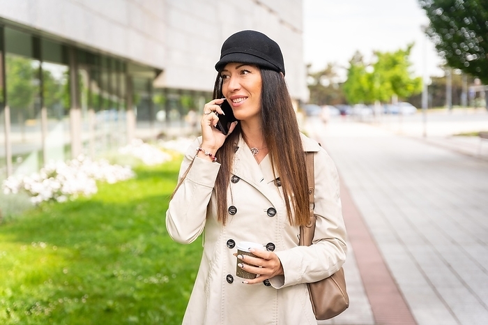 Businesswoman with a coffee in her hand and smiling talking on the phone outside the office, wearing a trench coat