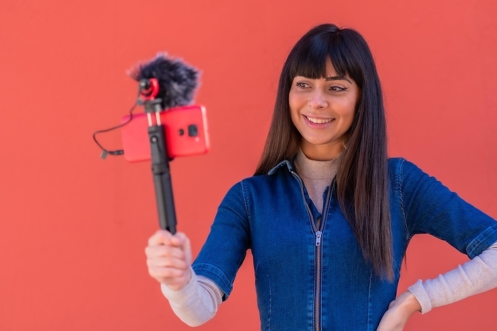 Brunette girl recording a video blog with the mobile phone in a blue denim outfit for social networks. Caucasian pretty a red background