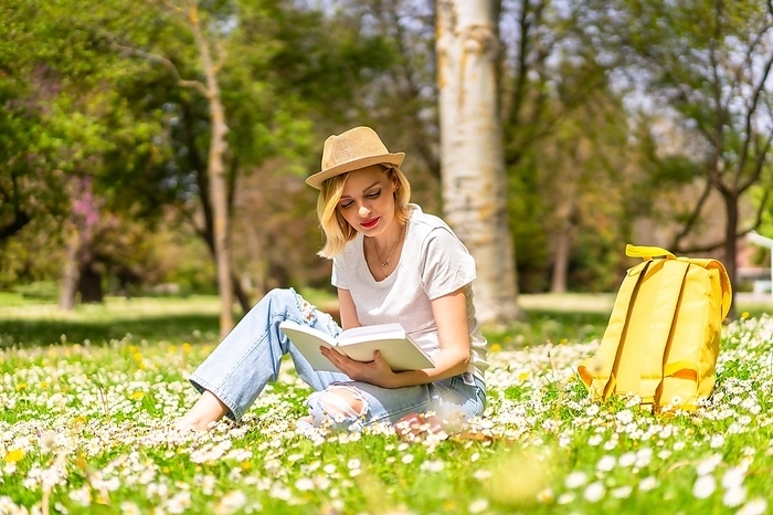 A young blonde girl in a hat reading a book in spring in a park in the city, vacations next to nature and next to daisies