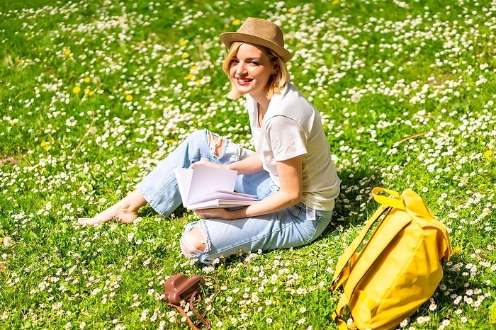 A young blonde girl in a hat writing poems in a notebook in spring in a park in the city, nature, sitting on the grass next to daisies