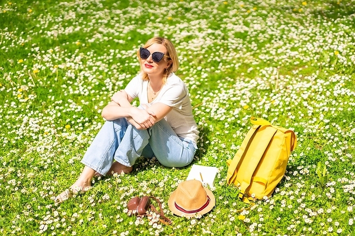 A young blonde girl in a hat unwinding and breathing pure haire in the spring in a park in the city, nature, sitting on the grass next to daisies