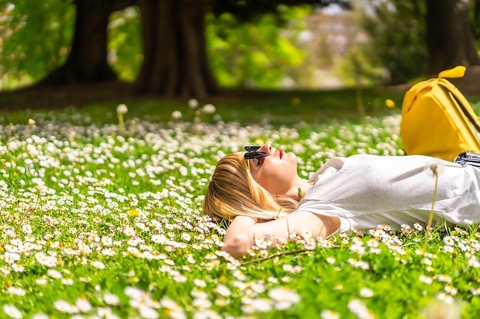 A young blonde girl in a hat unwinding and breathing pure hair in the spring in a park in the city, nature, lying on the grass next to daisies, looking at the camera