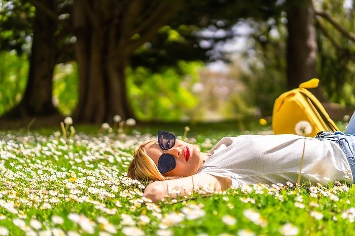 A young blonde girl in a hat unwinding and breathing pure hair in the spring in a park in the city, nature, lying on the grass next to daisies, looking at the camera