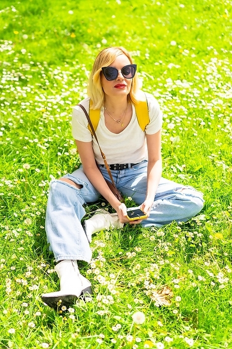 Portrait of a blonde tourist woman in a hat and sunglasses sitting on the grass in spring next to daisies, looking at the camera and enjoying nature