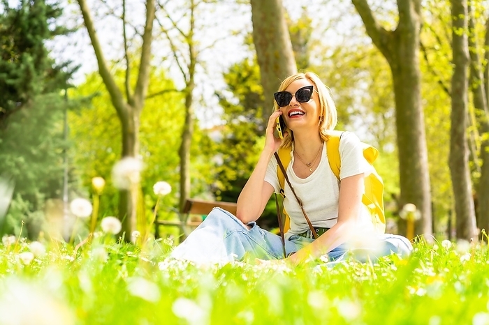 A smiling blonde tourist woman calling her friends with a phone, wearing a hat and sunglasses sitting on the grass in spring next to daisies in a park in the city