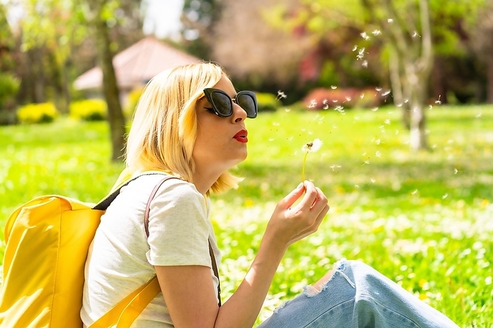 A tourist blonde woman blowing dandelion plant, wearing a hat and sunglasses sitting on the grass in spring next to daisies in a park in the city