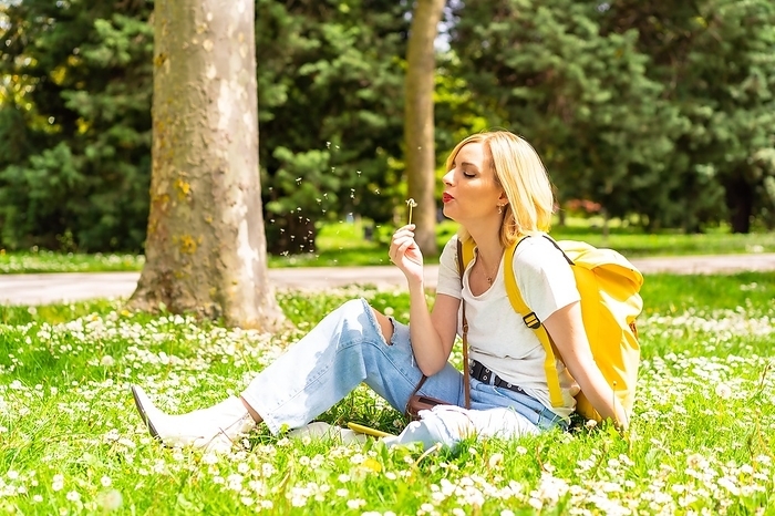A blonde woman blowing on the dandelion plant in a park in the city, wearing a hat and sunglasses sitting on the grass in spring next to daisies