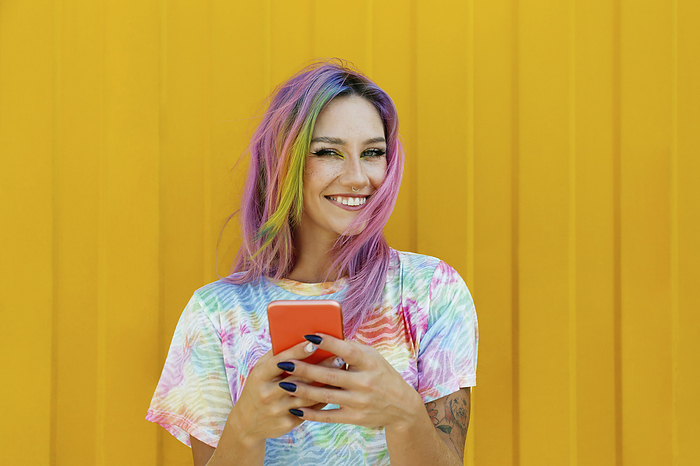 Happy young woman holding mobile phone in front of yellow wall
