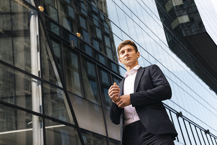 Contemplative young businessman adjusting cuff and standing in front of building