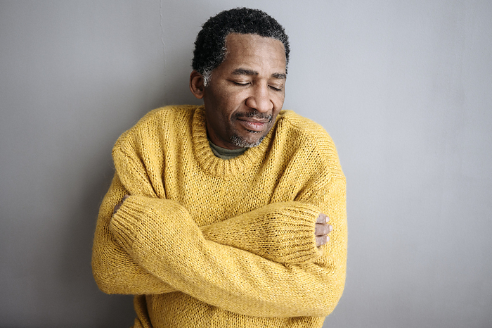 Mature man wearing yellow sweater with eyes closed leaning on gray wall