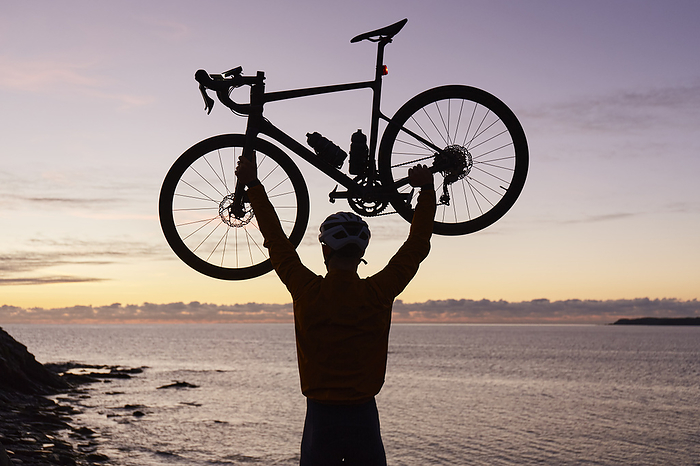 Cyclist carrying bicycle above head in front of sea at sunrise