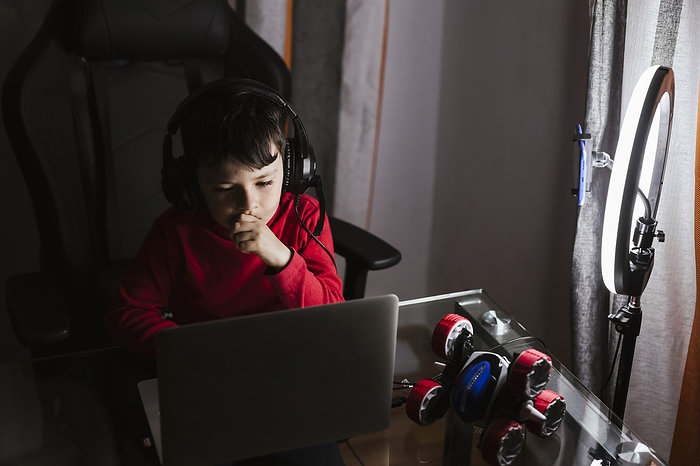 Boy wearing headphones using laptop sitting by mobile phone in illuminated ring light