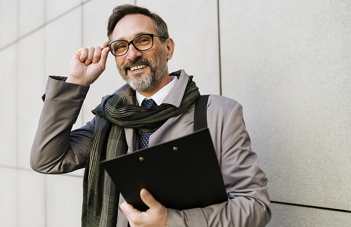 Happy mature businessman adjusting eyeglasses in front of wall