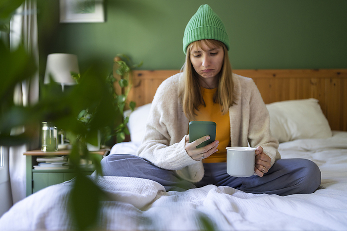 Sad woman using smart phone on bed with cup at home
