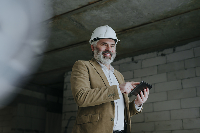 Happy businessman with hardhat standing with tablet PC at construction site