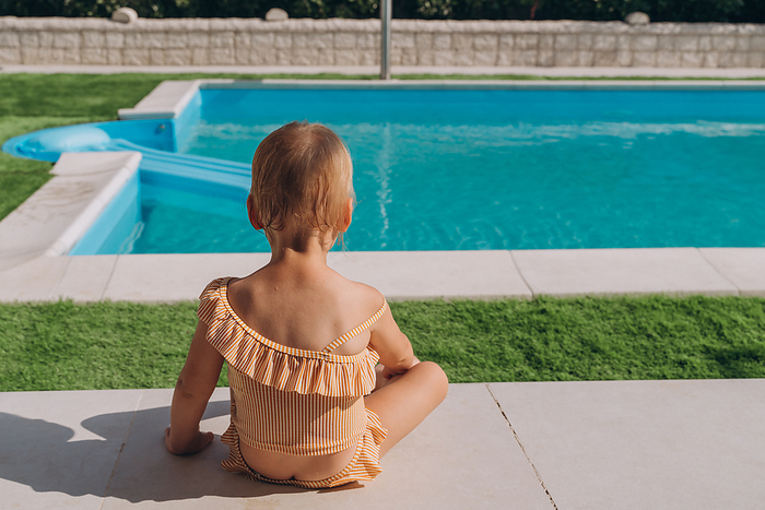 Baby in a bathing suit sits on a synthetic lawn near the outdoor pool