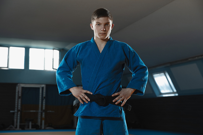 Young judo fighter in kimono posing comfident in the gym, strong and healthy Young judo caucasian fighter in blue kimono with black belt posing confident in the gym, strong and healthy. Practicing martial arts fighting skills. Overcoming, reaching target, self building up.