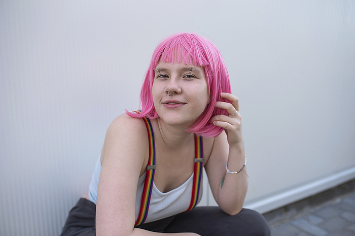 Smiling non-binary person with pink wig sitting in front of gray wall