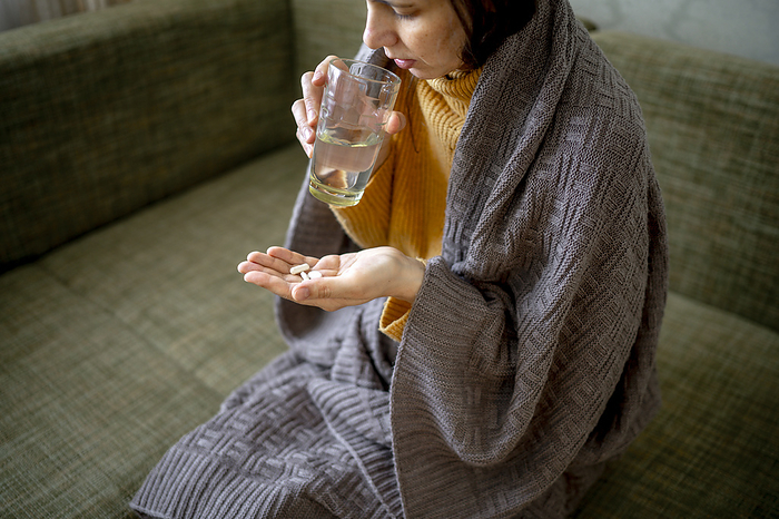 Sick woman wrapped in blanket taking medicine at home