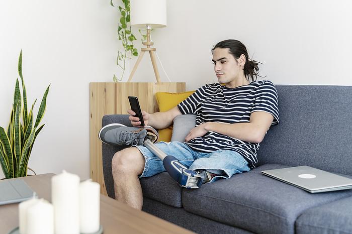 Young man with prosthetic leg using smart phone on sofa at home