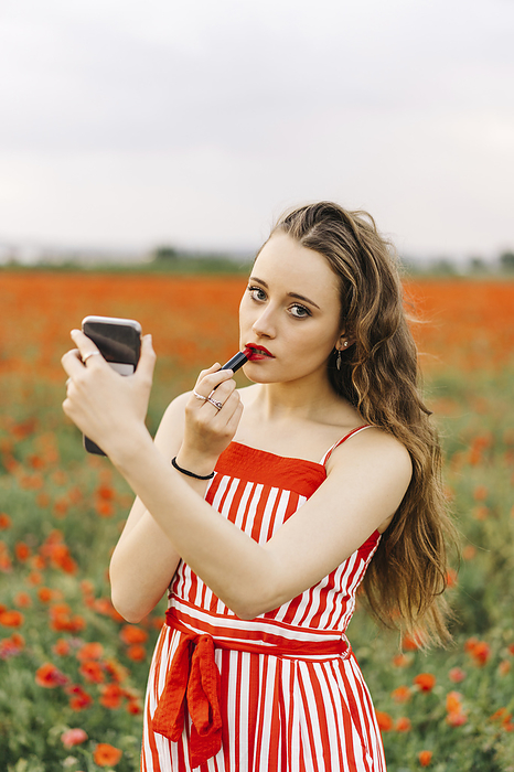 Young woman applying lipstick standing at poppy field