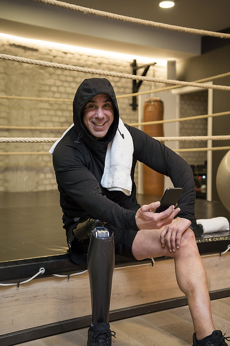 Happy man with disability sitting on boxing ring holding smart phone in gym
