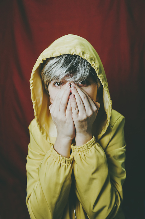 Woman wearing yellow hooded jacket covering mouth against red backdrop