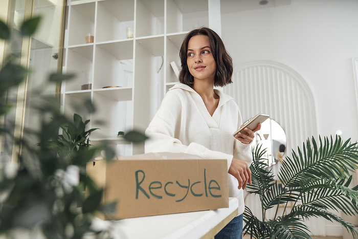 Smiling young woman with recycle box and smart phone standing at home