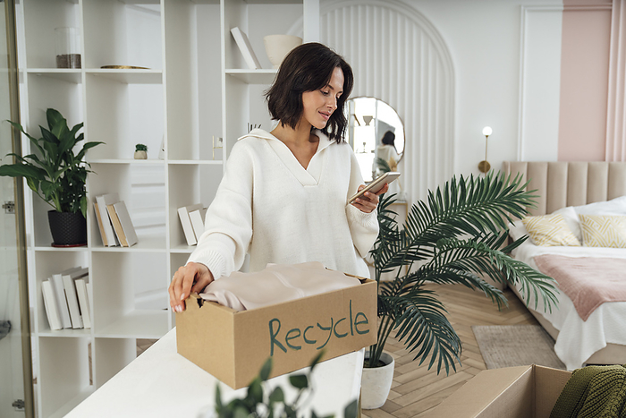 Smiling woman holding recycle box and using mobile phone at home