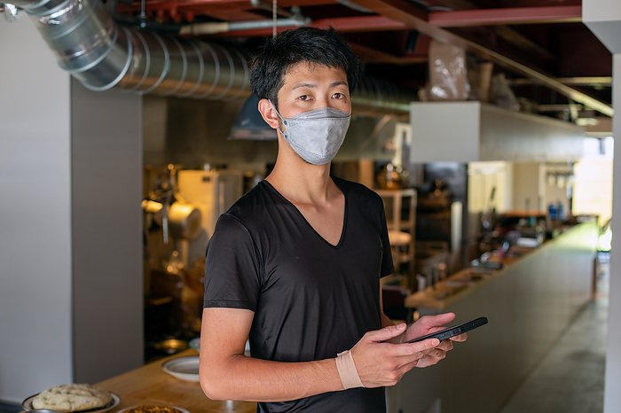 Restaurant,Japan,A man working in a restaurant, wearing a face mask, by an open kitchen, holding a digital tablet. A man working in a restaurant, wearing a face mask, by an open kitchen, holding a digital tablet.