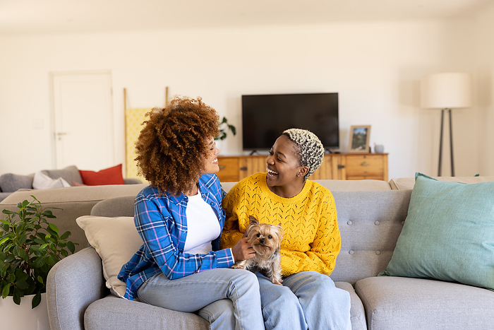 Cheerful diverse lesbian couple with cairn terrier relaxing on sofa in living room, copy space. Unaltered, dog, pet, care, love, togetherness, homosexual, afro hair, short hair and home concept.