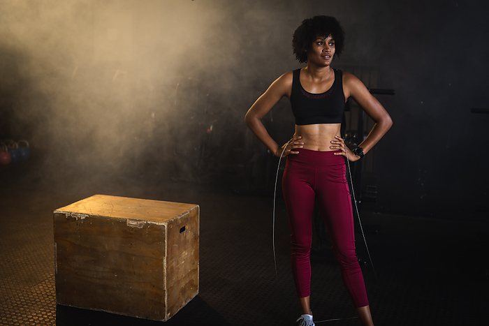 Tired biracial young woman with hands on hip holding jumping rope and standing in gym, copy space. Unaltered, cross training, exercise, afro hair, box, fitness and healthy lifestyle concept.