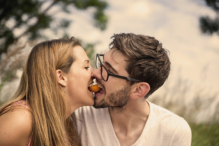 Mid adult couple biting peach together in the countryside, Bavaria, Germany Mid adult couple biting peach together in the countryside, Bavaria, Germany