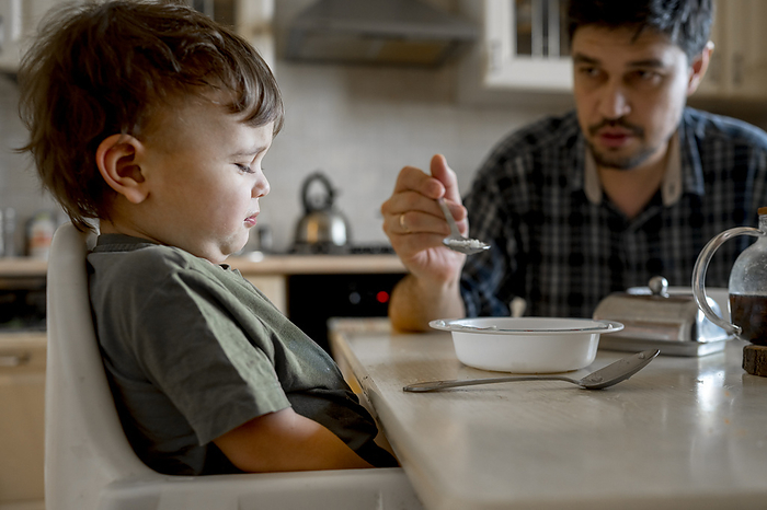 Father feeding upset son sitting at dining table in home