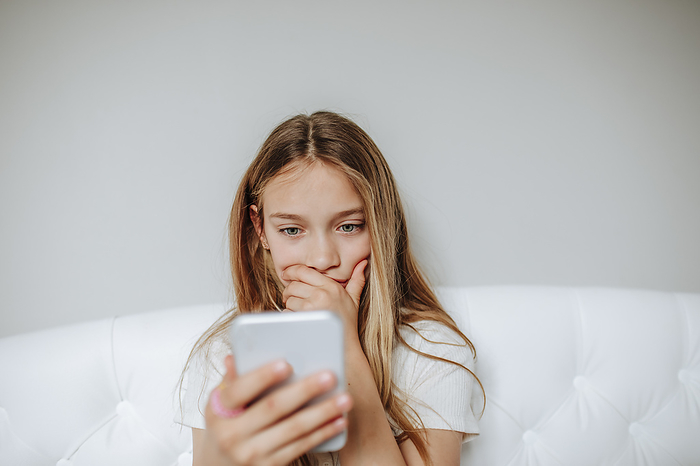 Worried girl using smart phone sitting on bed at home