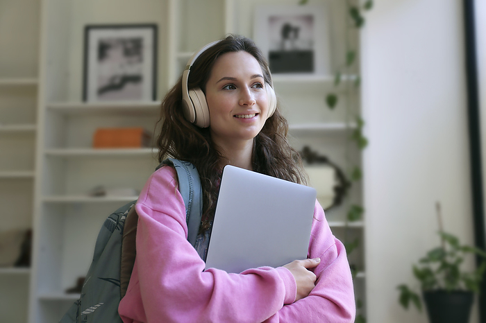 Smiling student wearing wireless headphones standing with laptop at cafe