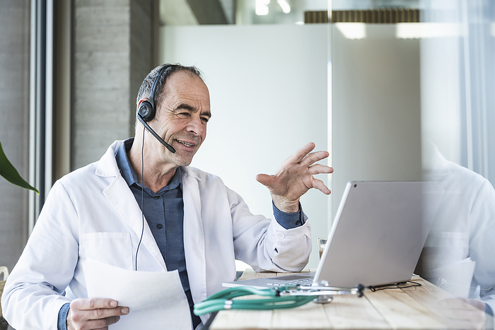 Smiling mature doctor explaining over telemedicine video call at desk
