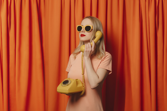 Woman wearing sunglasses talking over landline phone in front of orange curtains