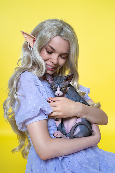 Young female cosplay elf in blue dress smiles happily, gently hugging Sphinx kitten to her chest Young female cosplay elf in blue dress smiles happily, gently hugging Sphinx kitten to her chest, by Zoonar Alexander A.