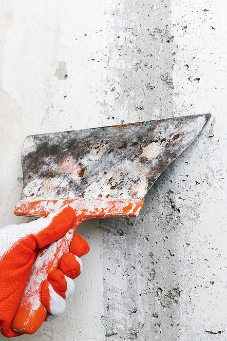 Hand of plasterer holds old flexible blade spackle knife and scrapes concrete wall, removes plaster, by Zoonar/Alexander A.