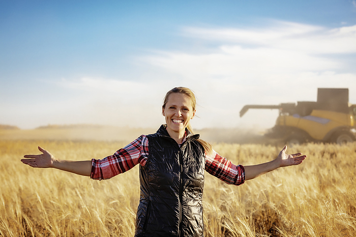 Portrait of a mature farm woman standing in a grain field with arms outstretched, posing for the camera during harvest with a combine working in the background at sunset; Alcomdale, Alberta, Canada, by LJM Photo / Design Pics