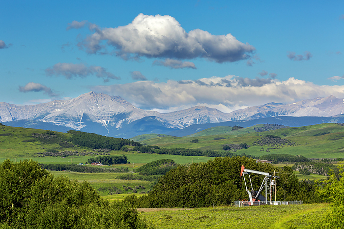 Canada Pumpjack in a green field with trees, rolling hills, mountain range, blue sky and clouds in the background, North of Longview, Alberta  Alberta, Canada, by Michael Interisano   Design Pics