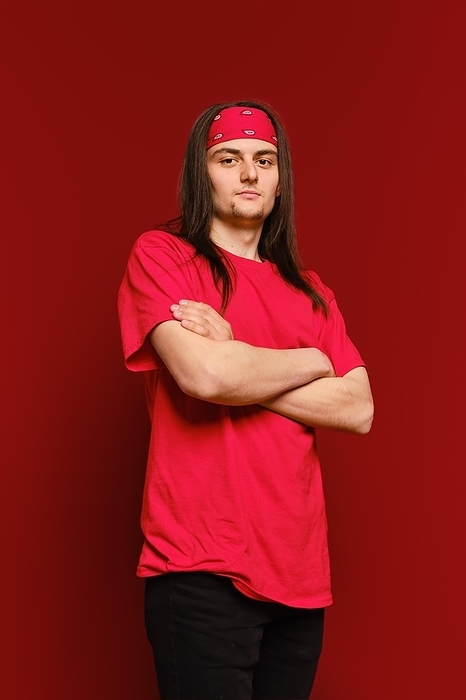 A young adult man in red shirt and head band stands in a studio with arms crossed and looks confidently at the camera against red background, by Aleksei Isachenko