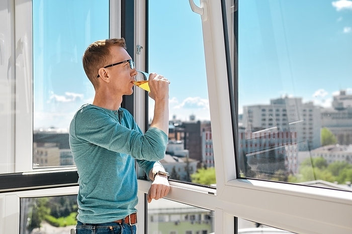 Middle aged man stands on balcony of high-rise building leaning against window frame and drinking juice, by Aleksei Isachenko