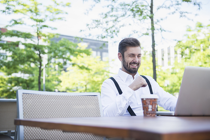 Businessman working on laptop in outdoor cafe and smiling, Munich, Bavaria, Germany Businessman working on laptop in outdoor cafe and smiling, Munich, Bavaria, Germany