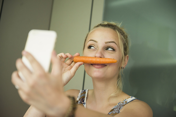 Young woman making a selfie with a carrot as a mustache Young woman taking a selfie and making moustache with a carrot on her face, Munich, Bavaria, Germany
