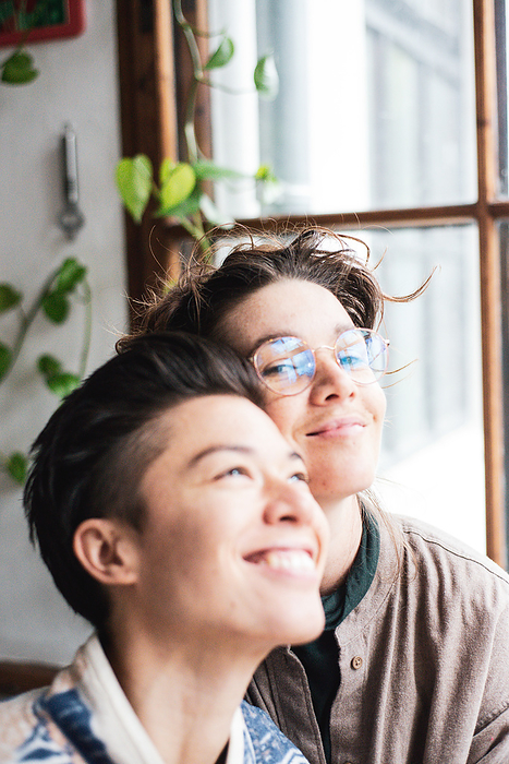 cute queer female couple smile as they cuddle by window at home